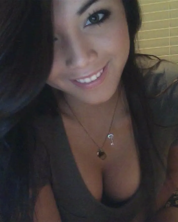Femme asiatique aux cheveux bruns noirs, souriante, portant un haut gris transparent décolleté, dans une chambre avec des stores.