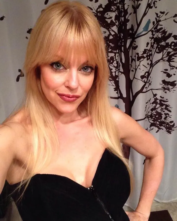Femme aux cheveux blonds, en robe noire sans bretelles, posant devant un rideau imprimé d'arbres.