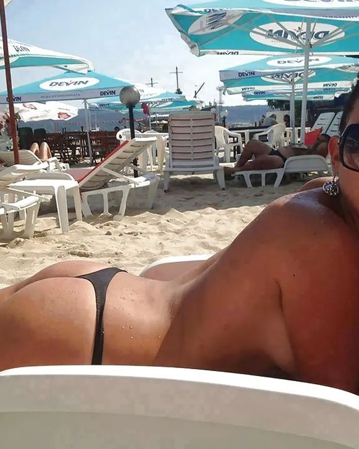 Femme aux cheveux bruns-noirs, en maillot de bain beige, posant sur une plage, souriante.