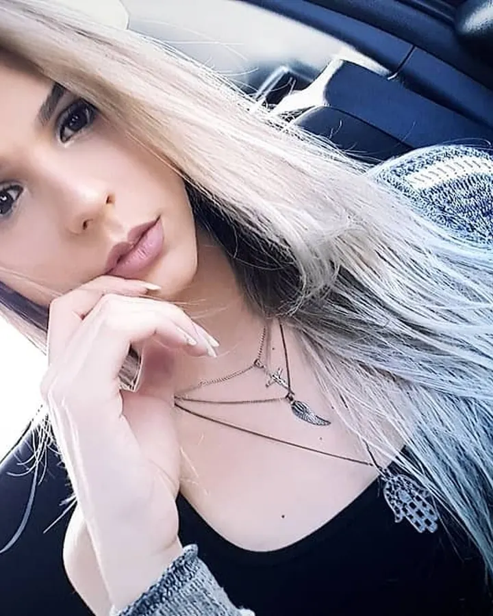 Femme aux cheveux platine assise dans une voiture, regardant l'appareil, vêtue d'un haut noir et d'un pull tricoté bleu.