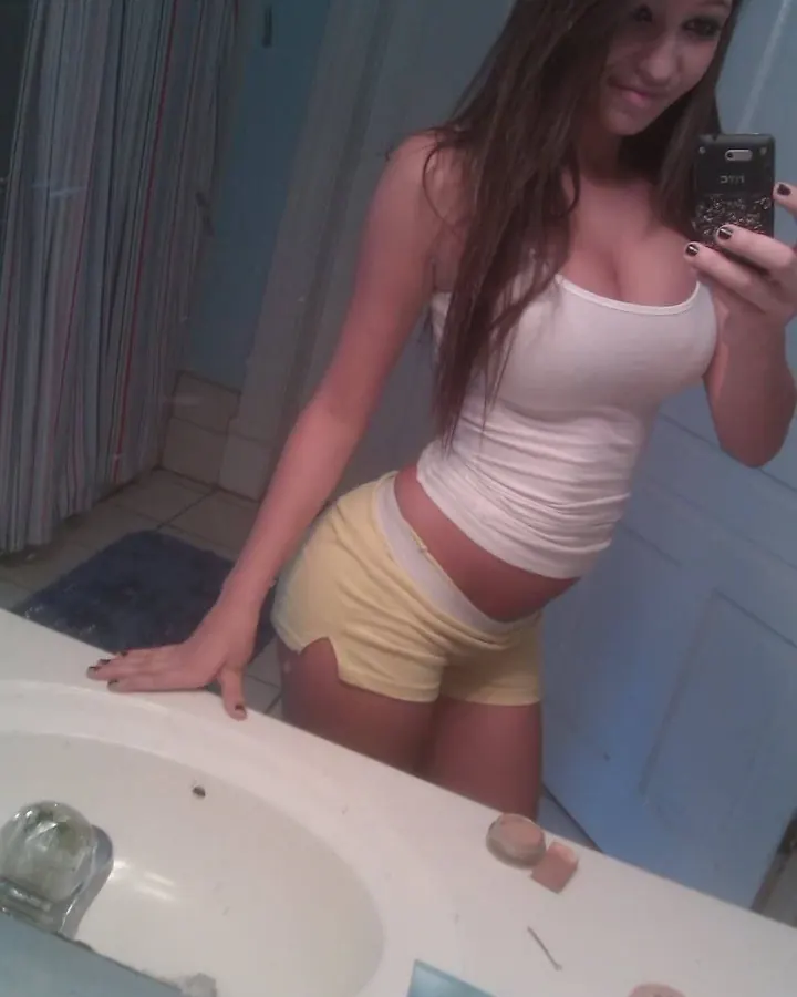 Jeune femme aux cheveux bruns, en haut blanc et shorts jaunes, prenant un selfie devant un lavabo dans une salle de bain.