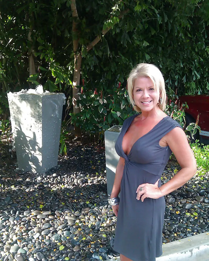 Femme blonde en robe grise, souriante, posant dans un jardin avec des plantes vertes