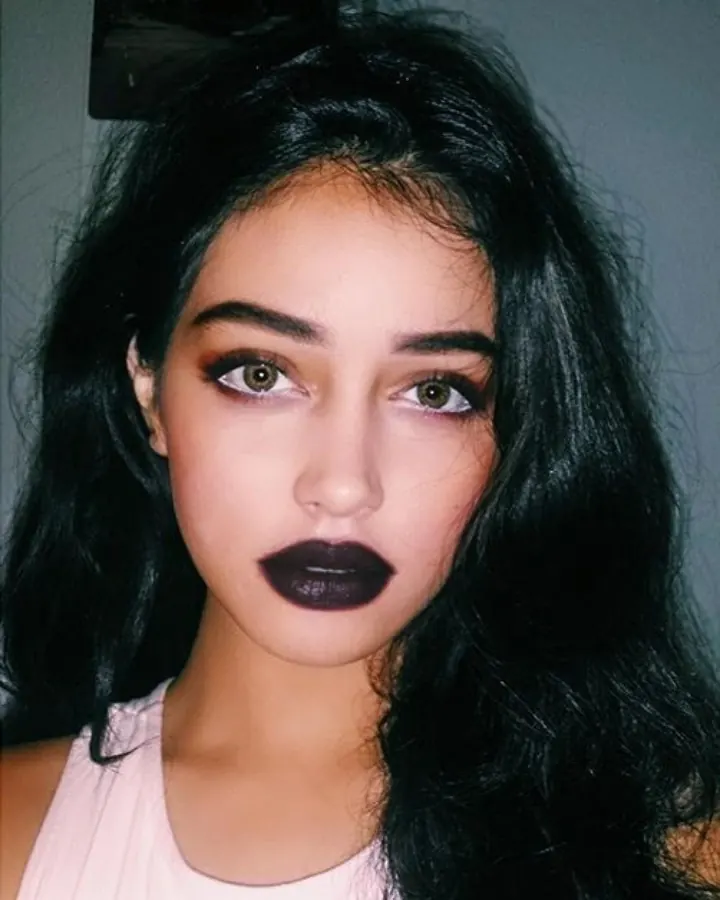 Femme latino aux cheveux bruns-noirs, en haut noir, regardant directement l'appareil photo avec un maquillage sombre et des lèvres foncées