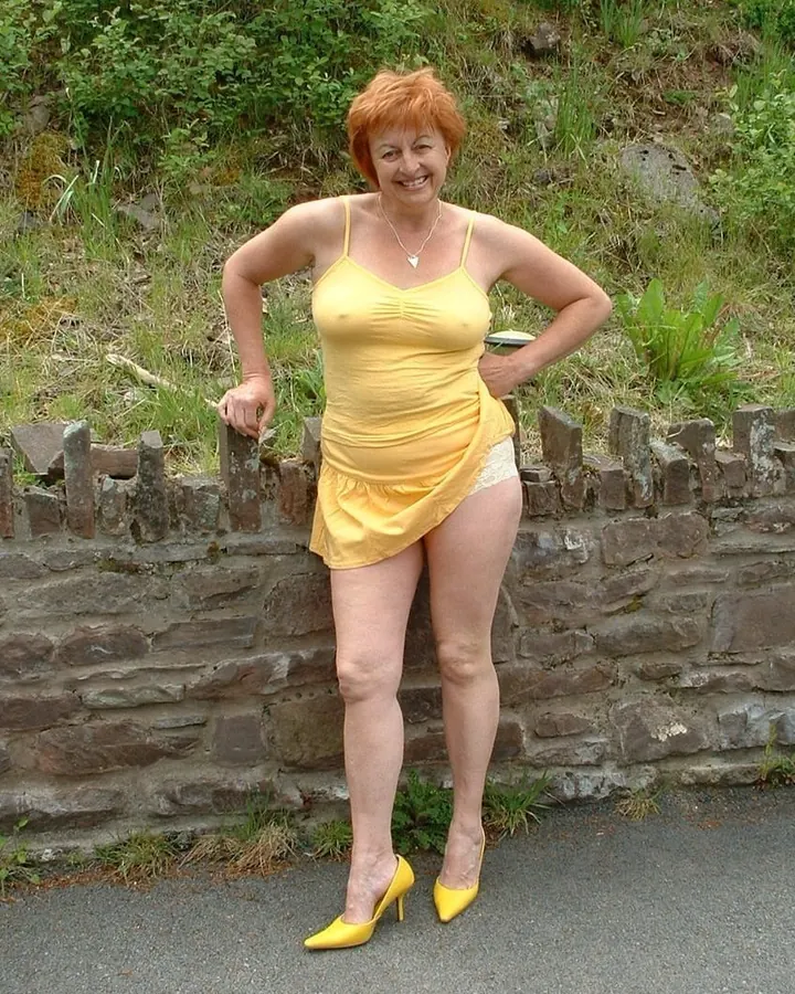 Femme blonde aux cheveux roux, en robe jaune, souriante, posant contre un mur de pierre