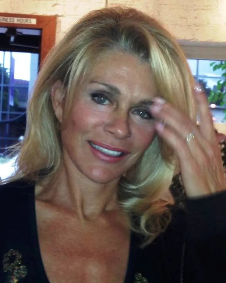 Femme blonde d'âge mûr, souriante, touchant son front avec une main, vêtue d'un haut noir