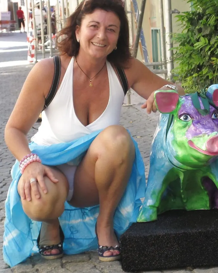 Femme aux cheveux bruns, souriante, assise sur un trottoir, portant un haut blanc et une jupe bleue, posant près d'une statue colorée.