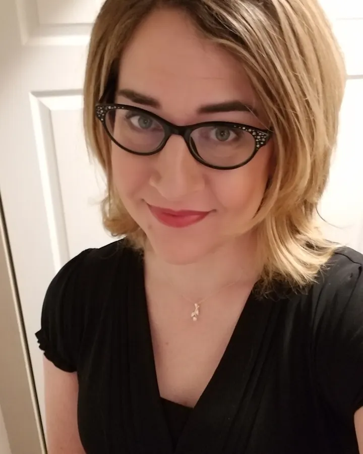 Femme aux cheveux blonds courts, lunettes noires ornées, souriant légèrement, vêtue d'une robe noire.