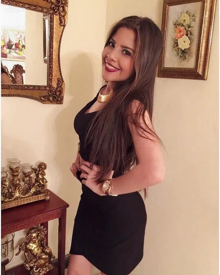 Femme aux cheveux bruns, souriante, vêtue d'une robe noire, posant près d'un miroir doré et d'une table décorative.