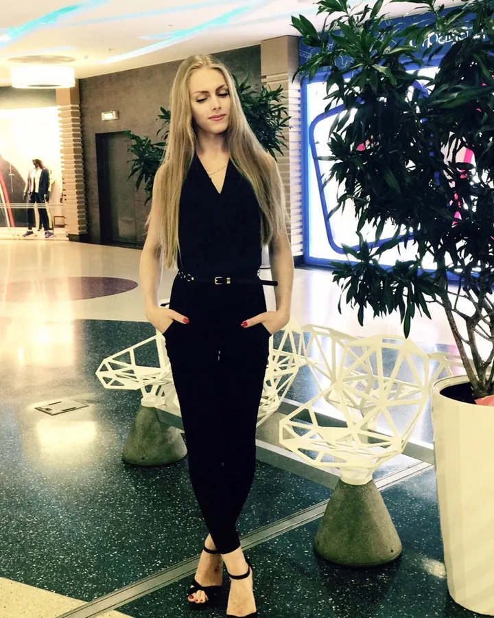 Femme blonde, vêtue d'une combinaison noire, mains dans les poches, regardant vers le bas dans un hall moderne.