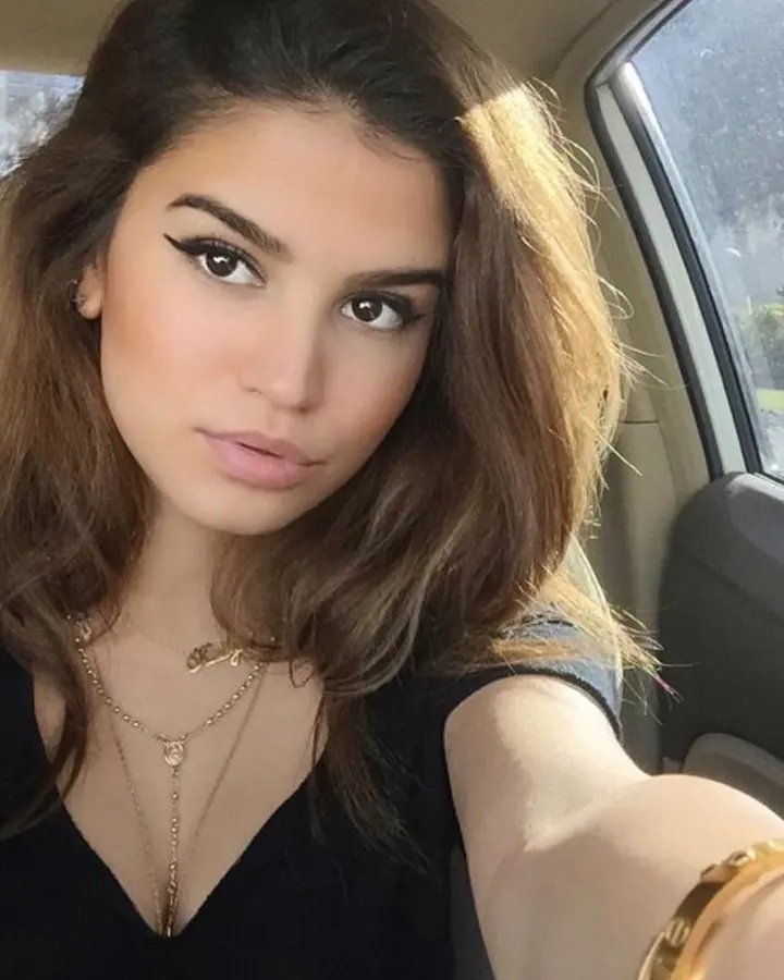 Jeune femme aux cheveux bruns, assise dans une voiture, regardant l'appareil photo, portant un haut noir et des bijoux dorés.