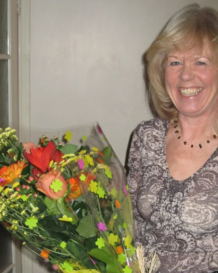 Femme blonde, en robe rouge, souriante, tenant un bouquet de fleurs colorées, dans une ambiance intérieure.