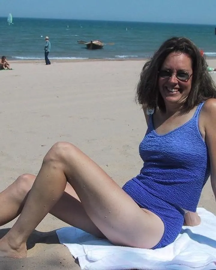 Femme aux cheveux bruns, souriante, en maillot de bain bleu, assise sur une plage, regardant l'appareil.