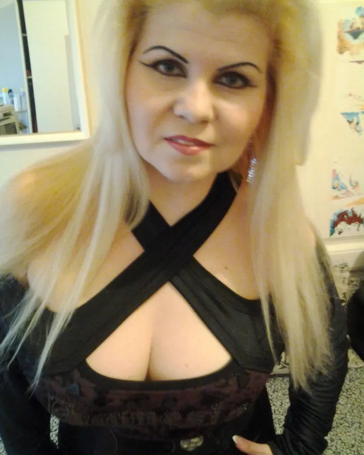 Femme aux cheveux platine, maquillage prononcé, vêtue d'un haut noir croisé, regardant l'appareil photo.