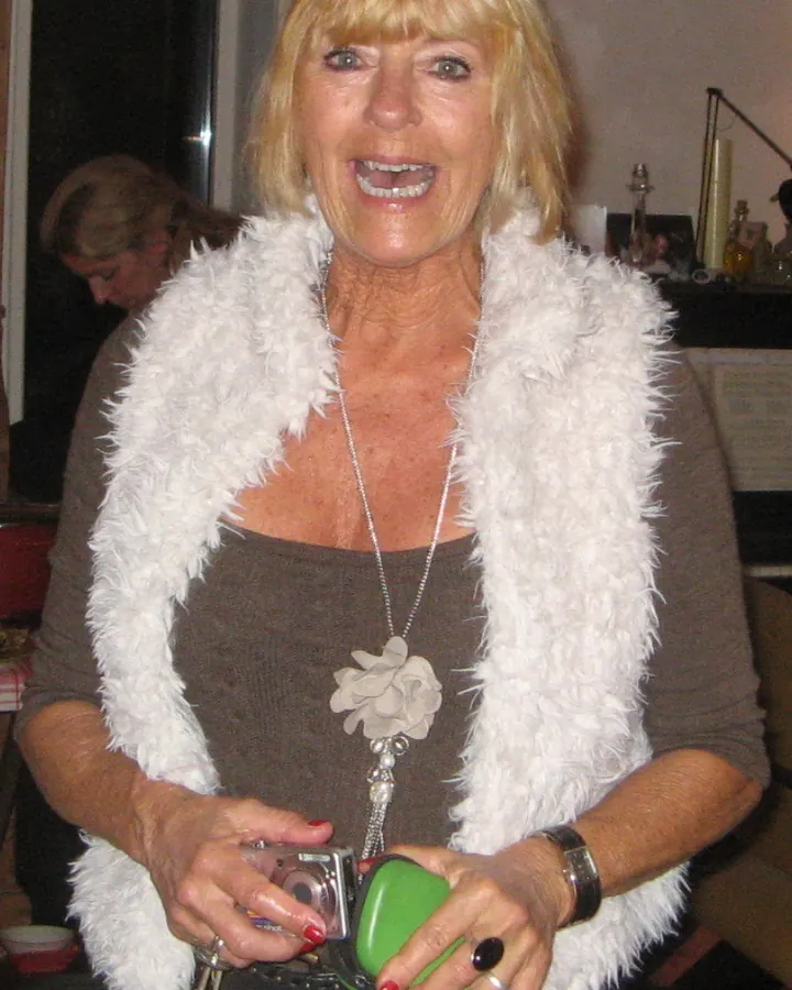 Femme blonde, souriante, portant une veste en fausse fourrure blanche, tenant un appareil photo et un objet vert.