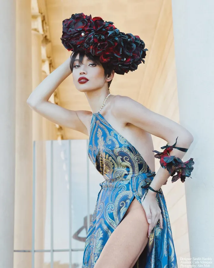 Femme asiatique en robe bleue ornée de motifs dorés, coiffée d'un chapeau de fleurs noires et rouges, posant près d'une colonne dans un cadre lumineux.