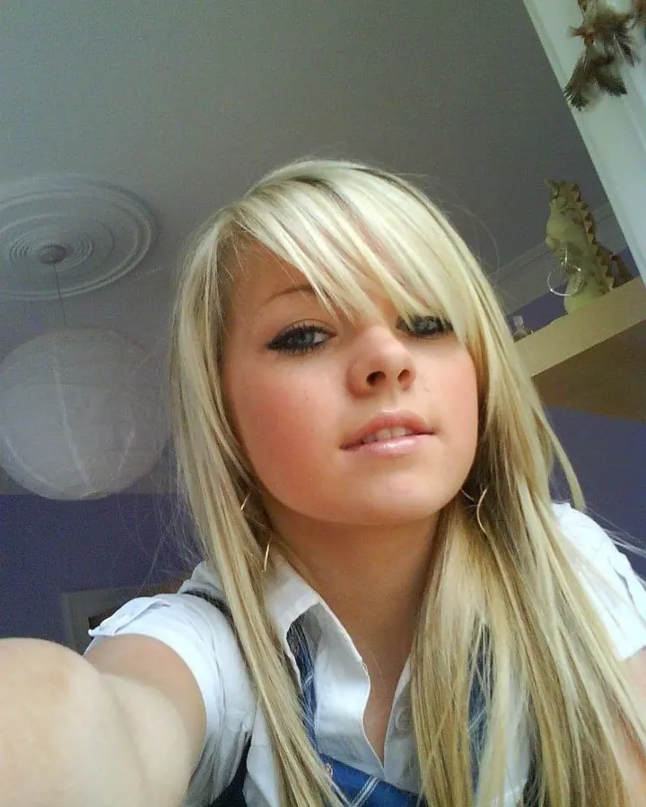 Jeune femme blonde en chemise blanche et tablier bleu, prenant un selfie dans une chambre aux murs violets.