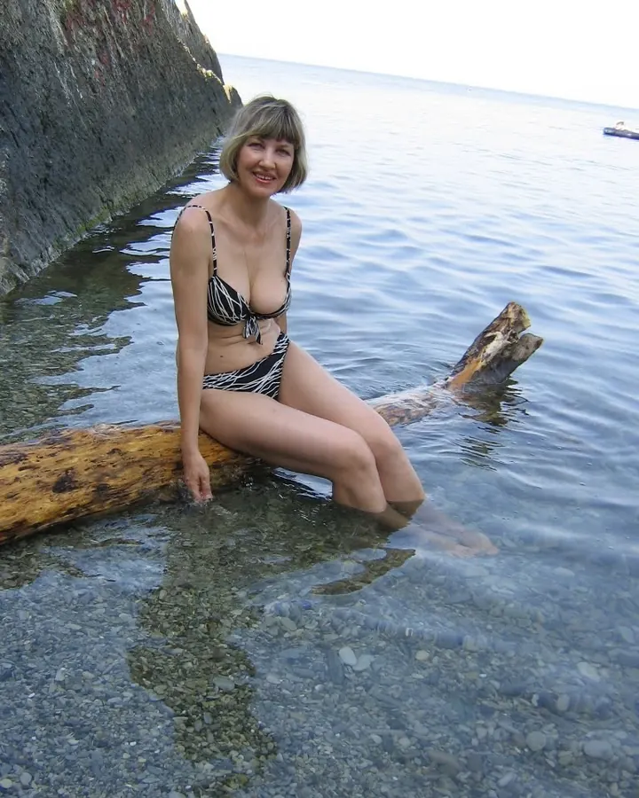 Femme blonde souriante, en maillot de bain noir et blanc, assise sur un tronc d'arbre dans l'eau claire près d'une roche.