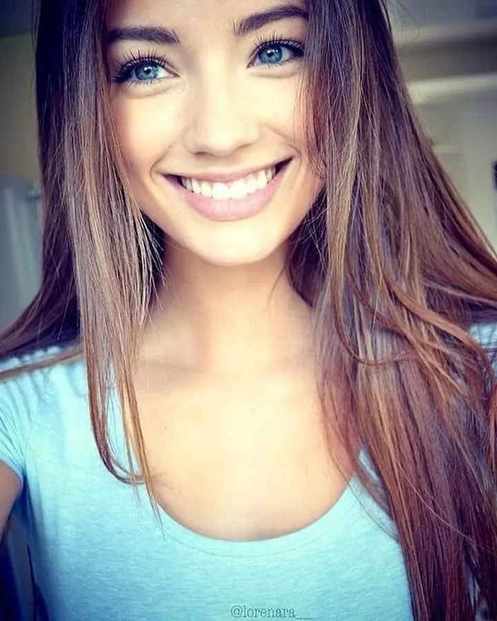 Jeune femme aux cheveux bruns, vêtue d'un haut bleu clair, souriant largement à la caméra dans une pièce intérieure.